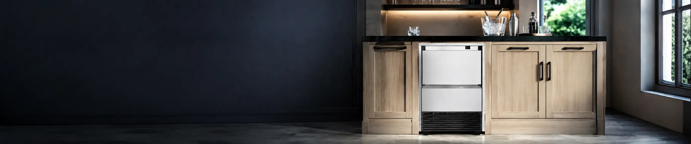 A stainless steel undercounter ice machine with a black front grille and feet, installed in a modern kitchen cabinet with light wood drawers and a dark countertop.slider_item_MKhKeX