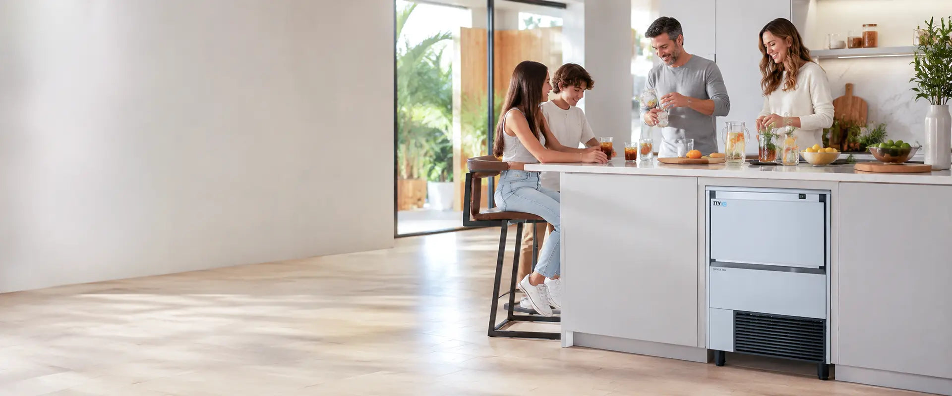 A modern kitchen scene with four people gathered around a white island counter. A compact white and black countertop ice machine is visible beneath the counter.1621243260e1af0c20-0